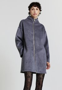 Donna con i capelli corti e ricci, che indossa una giacca grigia oversized con zip e collant neri con motivo in pizzo, in piedi davanti a uno sfondo semplice.