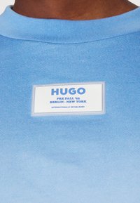 Camisa de manga corta azul claro con cuello acanalado, que presenta una etiqueta rectangular blanca con texto azul que dice "HUGO" y detalles adicionales debajo.