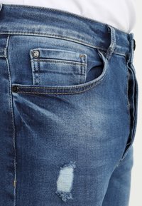 Blå denimjeans med slitage detaljer på vänster lår, kontrasterande sömmar och en liten myntficka på högra sidan.