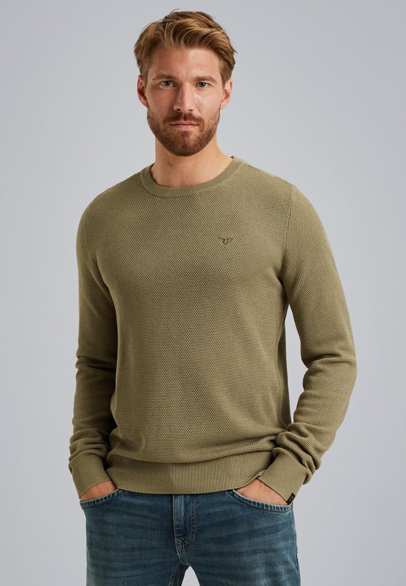 Olivengrüner Strukturpullover mit Rundhalsausschnitt, schmalen Bündchen und kleinem Logo auf der Brust. Getragen mit blauen Jeans.