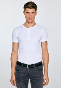 Fiatal férfi, aki szűk fehér, rövid ujjú henley pólót és fekete övet visel sötétszürke farmerrel, világosszürke háttér előtt áll.