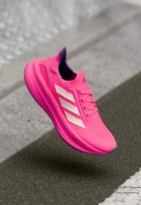 Rosa Sportschuh mit strukturiertem Obermaterial, ausgestattet mit drei weißen Streifen, einer gepolsterten Sohle und einem lila Akzent am Schaft.