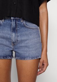 Osoba v modrých denimových kraťasech s mírným pasem a černém krátkém tričku, s pravou rukou uvolněně položenou na boku proti jednobarevnému pozadí.