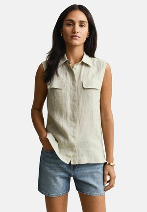 Woman with long dark hair wearing sleeveless beige button-up shirt and blue denim shorts, standing with one hand in pocket.