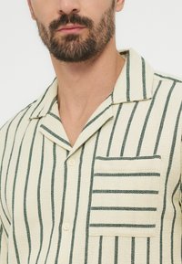 Άνδρας με περιποιημένο γένι, που φοράει κρεμ πουκάμισο με κατακόρυφες σκούρες πράσινες ρίγες και τσέπη στο στήθος με οριζόντιες ρίγες.