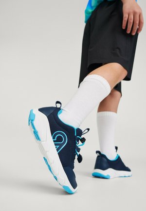 Persona che indossa calzini bianchi alla caviglia, pantaloncini neri e scarpe da ginnastica blu navy con accenti azzurri, alzando un piede a metà passo su uno sfondo neutro.