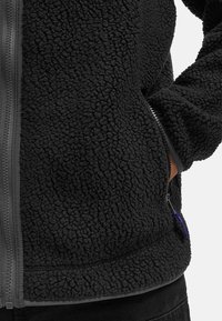 Giacca in pile nera con una superficie testurizzata e accogliente. Presenta una chiusura frontale con zip e tasche laterali con un tirante della zip di colore viola a contrasto.