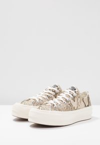 Sneakers platform basse con motivo a stampa di serpente beige e marrone e lacci bianchi su una superficie bianca contro uno sfondo neutro.