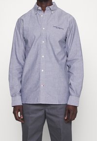 Vaaleansininen chambray-kankainen napillinen paita, jossa on kaulus, pitkät hihat, valkoiset napit ja pieni logo-ristikon yksityiskohta rintataskussa.