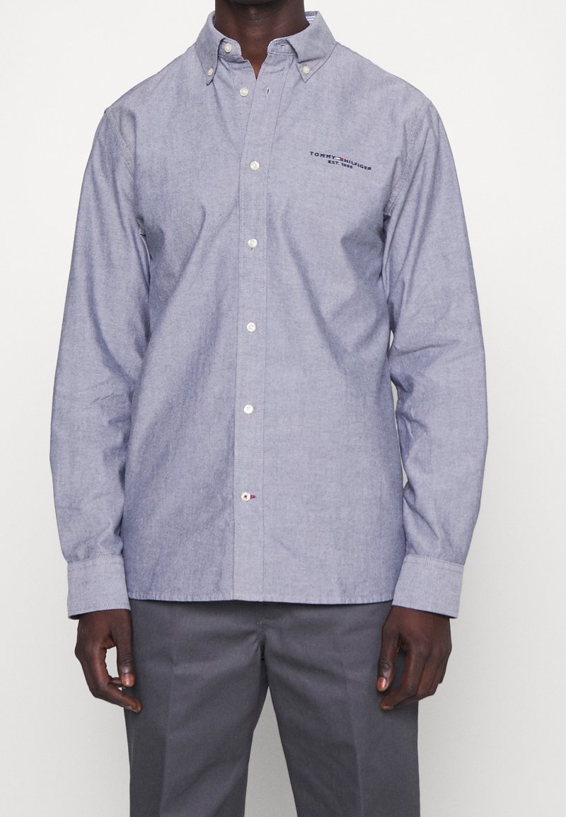 Vaaleansininen chambray-kankainen napillinen paita, jossa on kaulus, pitkät hihat, valkoiset napit ja pieni logo-ristikon yksityiskohta rintataskussa.