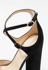 Černé semišové sandály na vysokém podpatku s kotníkovými pásky a stříbrnými přezkami. Mají široký podpatek a hladovou béžovou stélku se značkovým logem.