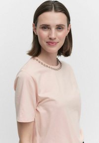 Ljust rosa t-shirt med kort ärm och rund halsringning prydd med en kedjeformad dekoration. Mjuk tyg och enkel design.