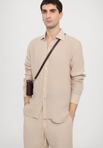 Camisa de linho bege com botões, mangas longas e um hem em curva, combinada com calças a condizer. Uma pequena bolsa de couro borgonha pende de uma alça.