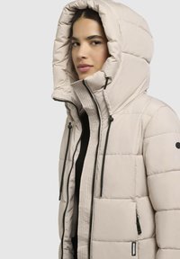 Beige puffer jakk, millel on kõrge, tikitud kapuuts, tõmblukuga esiosa ja mustad tõmblukud. Omab musta logo plaastrit ja tekstureeritud külgpaneele.