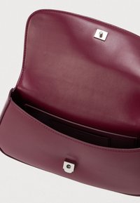 Burgundi värvi nahast kott avatud klapiga, hõbedane lukustus ja sise-tasku heleda tausta ees.