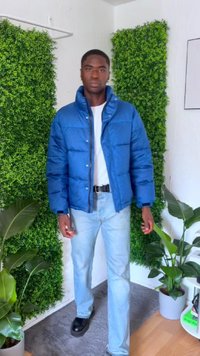 Modrá páperová bunda s prešívanou textúrou, predným zipsom a vysokým golierom; skombinovaná s bielou košeľou a svetlomodrými džínsami. Jednoduché čierne topánky.