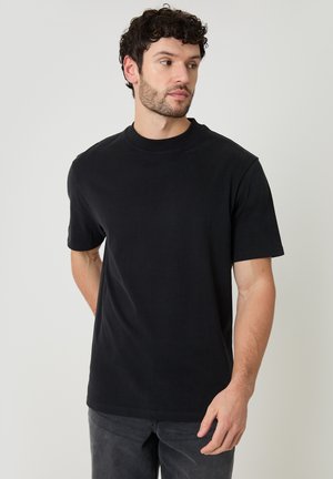 Man met krullend haar die een effen zwart T-shirt met korte mouwen draagt, staand tegen een lichtgrijze achtergrond.