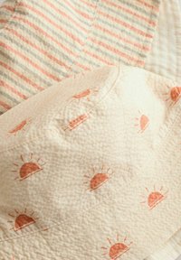 Crème emmerhoed met zonnenpatroon in oranje. Heeft een gestructureerde stof en een gestreepte achterzijde in lichtroze, blauw en crèmekleuren.