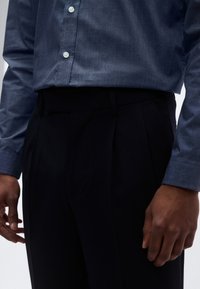 Mėlyna marškiniai su tekstūruota medžiaga, dėvimi su tamsiomis plisuotomis kelnėmis. Matoma ranka, poilsiaujanti ant šlaunies, kuri pabrėžia aprangos pritaikymą.