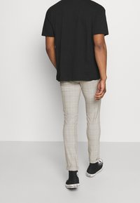 Svart kortärmad skjorta, ljusa rutiga byxor i beige nyanser med grå linjer, och svarta sneakers med vita sulor. Bakifrån.