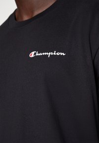 Sort bomulds t-shirt med et hvidt "Champion" logo på venstre side. Stoffet har en glat, blød tekstur og klassisk crew neck design.