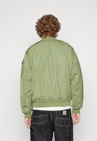 Giubbotto bomber verde oliva con zip frontale, colletto e polsini a coste e finitura testurizzata. Presenta tasche sulle spalle.