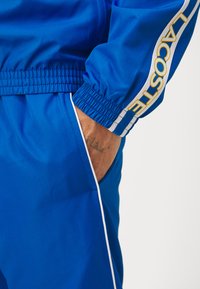 Kék sportos melegítő, texturált anyaggal, rugalmas mandzsettákkal, és sárga "LACOSTE" logóval az ujjon. Fehér díszítéssel.