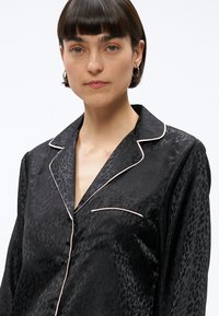 Młoda kobieta z krótkimi, ciemnymi włosami nosi czarną satynową, panterkową piżamę z jasnoróżowym obszyciem i małą kieszonką.