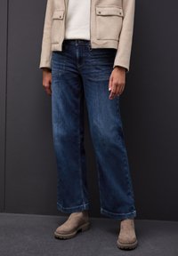 Mörkblå vida jeans i denim, med en medelhavs tvätt och kontrasterande sömmar, kombinerat med en beige mockajacka och bruna ankelstövlar.