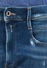 Tumed sinine denim, millel on kulunud tekstuur, esipocketid ja kontrastsed kollased õmblused. Metallne nööp sulgemiseks vööl.