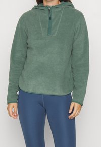 Pullover in pile verde con cappuccio, zip a quarto, superficie texturizzata e orlo curvo. Abbinato a leggings blu.