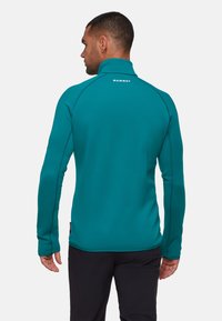 Man die met zijn rug naar de camera kijkt, gekleed in een teal Mammut-jas en zwarte broek, tegen een effen lichte achtergrond.