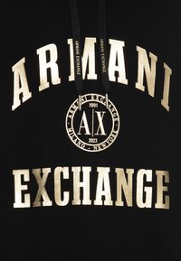 Sudadera negra con texto en dorado en negrita que dice "ARMANI EXCHANGE", logotipo circular y cordones. El material parece suave; el diseño es simple pero llamativo.