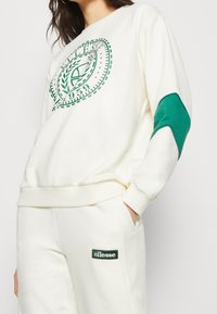 Felpa crema con stampa del logo verde e dettagli verdi sulle maniche. Abbinata a pantaloni jogger crema con un patch del logo verde.