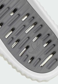 Grå gummisula med urtagna spår, vit accenttrim och en liten präglad logotyp. Slät yta med en texturerad greppyta.