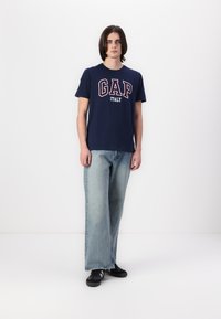 Tamno plava pamučna majica s natpisom "GAP ITALY" ispisanim crvenom i bijelom bojom. Uparena s svijetloplavim širokim trapericama i crnim tenisicama.