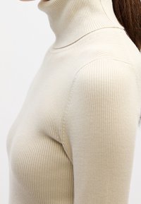 Gros plan d'une personne portant un pull à col roulé côtelé beige à manches longues, visible du cou au haut du torse.