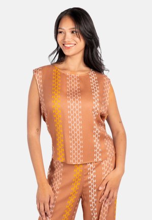 Kvinde smiler, iført brun ærmeløs top og matchende bukser med lodrette hvide og gule kæde-link mønstre.