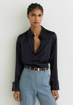 Ung kvinde med flettet hår iført en sort satinbluse stoppet ned i højtaljede blå bukser med påsyede lommer og et brunt bælte.