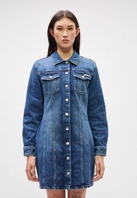 Μπλε φούστα από τζιν, με μακριά μανίκια, δύο τσέπες στο στήθος, κουμπιά μπροστά και εμφανείς λεπτομέρειες ραφής. Λείο υλικό, προσαρμοσμένη εφαρμογή.