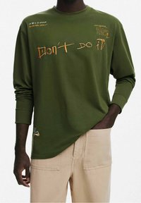 Maglietta a maniche lunghe verde oliva con testo grafico arancione, caratterizzata da varie frasi scritte a mano e un piccolo design ricamato sull'orlo.