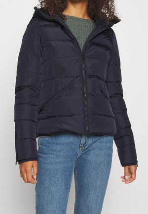 Femme portant une veste matelassée noire avec des poches zippées, un pull en tricot vert et un jean bleu, debout devant un fond clair uni.