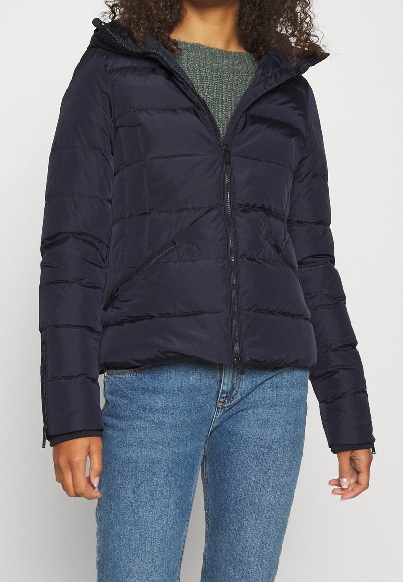 Femme portant une veste matelassée noire avec des poches zippées, un pull en tricot vert et un jean bleu, debout devant un fond clair uni.