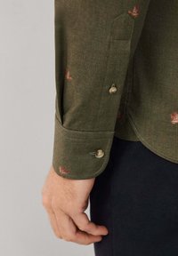 Mannenhand naast de lange mouw van een donkergroen shirt met kleine rode vogelprints en beige knopen, gecombineerd met zwarte broek.