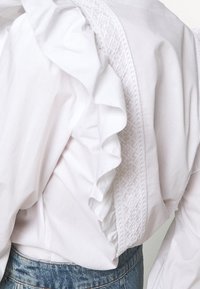 Blouse blanche avec des détails à volants et un panneau vertical brodé, portée avec un jean en denim bleu.