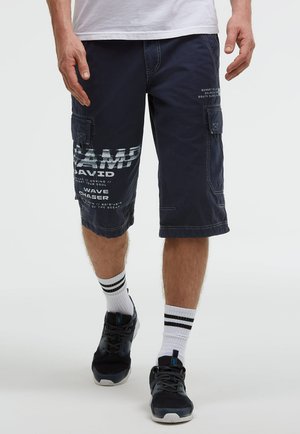 Mann trägt dunkelblaue Cargo-Shorts mit weißem Text, weiße Socken mit schwarzen Streifen und schwarze Sneakers, steht vor einem einfarbigen Hintergrund.