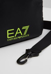 Róg czarnego paska torebki z neonowo zielonym logo EA7 Emporio Armani oraz czarną plastikową klamrą przymocowaną do czarnego plecionego paska na białym tle.