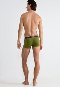 Green boxer shorts made of smooth fabric, featuring a black waistband with "sloggi" branding, shown from a rear view.
