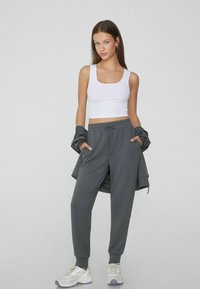 Weißes, kurzes Tanktop, graue Jogginghose mit Kordelzug und hellgraue Turnschuhe. Das Outfit vermittelt einen lässigen, sportlichen Stil.
