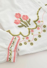 Valkoinen kangas, jossa on vaaleanpunainen tulppaanikirjailu, vihreä aaltoileva reuna ja koristeellisia vaaleanpunaisia ja vihreitä tikattuja kuvioita kalvosinnapin läheisyydessä.